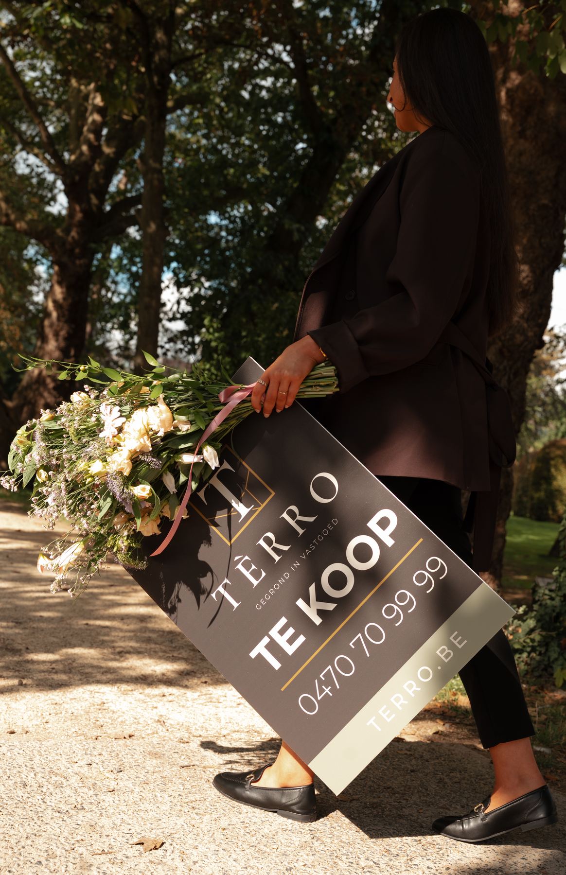Makelaar van TÈRRO wandelt door het park met bloemen en een bord Te koop